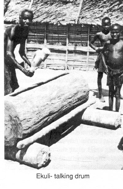 A man leaning over a large hollowed-log Ekuli talking drum while two children watch, thatched roofs behind